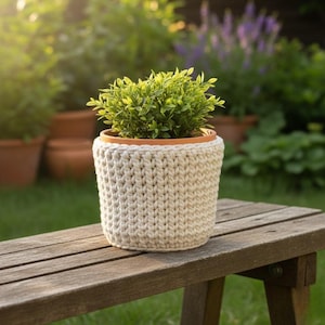 May include: A small potted plant with vibrant green leaves sits inside a cream-colored knitted cozy. The plant pot is made of terracotta. The cozy is on a wooden bench in a garden setting.