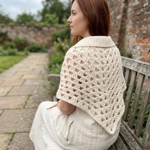 Peut inclure: Châle en crochet crème à tissage ouvert avec un col. Le châle est drapé sur les épaules. La personne est assise sur un banc en bois dans un cadre extérieur. Le châle est fait d'un fil doux et texturé.