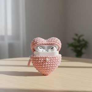 May include: A pink, heart-shaped crochet case for wireless earbuds. The case is open, revealing white earbuds inside. The case has a button closure and a small loop. The background is a light-colored wooden surface.