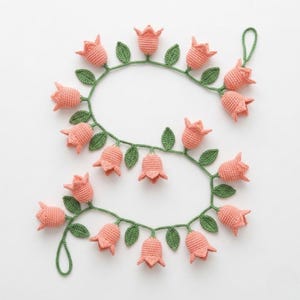 Op de afbeelding: Een gehaakte slinger met perzikroze tulpvormige bloemen en groene bladeren. De bloemen zijn gelijkmatig verdeeld over een groene rank, waardoor een decoratieve bloemenketting ontstaat. Geschikt voor woondecoratie.