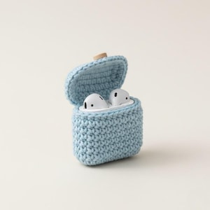 May include: A light blue, crocheted case for wireless earbuds, with the lid open to reveal the white earbuds inside. The case has a wooden button closure. The earbuds are white with black accents. The case is a soft, textured material.