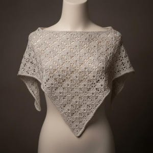 Può includere: Uno scialle all'uncinetto grigio chiaro e traforato, esposto su un manichino. Lo scialle presenta un motivo floreale e una forma triangolare. La scollatura è ampia, a barchetta. Il materiale appare morbido e leggero, adatto alla stratificazione.