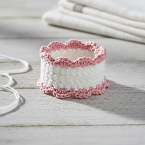 Könnte beinhalten: Ein handgefertigter Häkel-Serviettenring in Weiß mit einem rosa, gewellten Rand. Der Ring ist zylindrisch und steht auf einer hellen Holzoberfläche. Weiße Schnur ist im Vordergrund zu sehen, und gefaltete weiße Handtücher sind im Hintergrund.