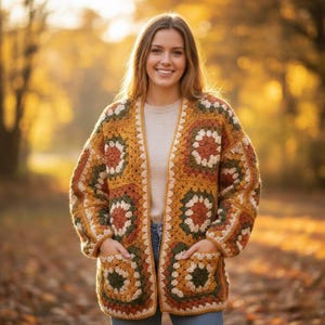 May include: A handmade, open-front crochet cardigan in a granny square pattern. The jacket features a color palette of mustard yellow, brown, green, and cream. It has two front pockets and is worn over a neutral-colored top and blue jeans.