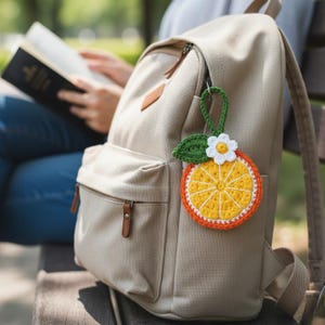 May include: A beige backpack with brown leather accents and a crocheted orange slice keychain. The keychain features a yellow and orange slice with a white flower and green leaf. The backpack is hanging on a person's back.