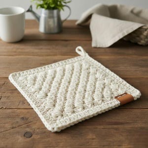 以下が含まれることがあります： クリーム色の、正方形のかぎ針編み鍋つかみ。テクスチャ加工された斜め模様が特徴です。小さな茶色の革タグと吊り下げ用のループが付いています。鍋つかみは木製の表面にあります。