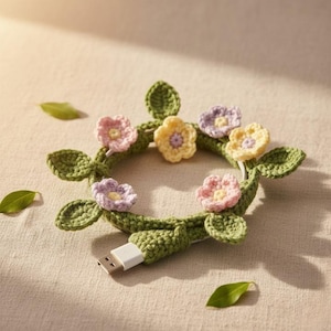 May include: A crocheted USB cable featuring a floral design. The cable is wrapped in green yarn, adorned with crocheted leaves and colourful flowers in pink, yellow, and purple. The USB connector is white.