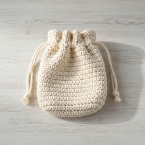以下が含まれることがあります： クリーム色の編み物巾着袋。バッグは太い糸でできており、底は丸みを帯びています。巾着紐は同じ糸でできており、端が結ばれています。