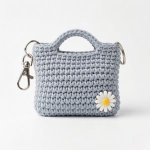 May include: A light blue crocheted mini tote bag with a handle and a daisy applique. The bag has a silver-colored clasp and a keyring attached. The bag is approximately 3 inches wide and 3 inches tall.