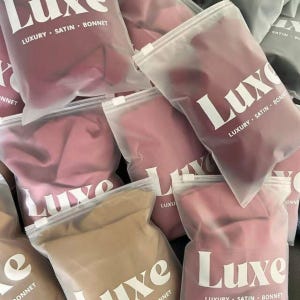Op de afbeelding: Een verzameling satijnen bonnets in verschillende roze en bruine tinten, verpakt in transparante plastic zakken met witte ritsen. De zakken zijn voorzien van het woord "Luxe" en de tekst "Luxury Satin Bonnet" in een vetgedrukte, witte letter.