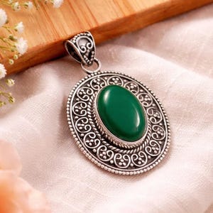 May include: A silver pendant with an oval, emerald-green gemstone at the center. The pendant has an ornate, circular design with a decorative bail. The jewelry is displayed on a white fabric surface.