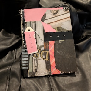 May include: A mixed media art journal with a pink tag that reads "hard core." The journal features torn paper, black and white images, and a black spine. The word "Truth" is repeated on a small tag. The journal is resting on a black leather surface.