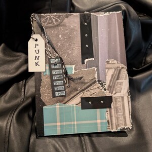 May include: A handmade journal with a collage design. The cover features torn paper, black accents, and a teal plaid pattern. A tag with the word "PUNK" hangs from the journal, along with a series of tags that say "Truth".