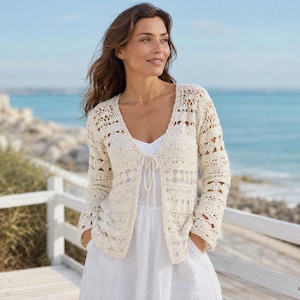 Könnte beinhalten: Cremefarbene, offene Häkeljacke mit langen Ärmeln und Bindeband. Das komplizierte, spitzenartige Design weist verschiedene Muster auf. Die Jacke wird über einem weißen Kleid getragen. Der Hintergrund zeigt eine Strandszene mit blauem Meer und einem weißen Holzzaun.