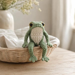May include: A handmade crochet frog with large, golden-rimmed eyes and a cream-colored belly sits in a woven basket. The frog is primarily green, with detailed crochet work. The frog is a whimsical decorative item.
