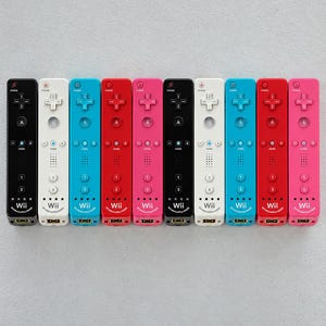 May include: A collection of ten Wii remote controllers arranged in a row, featuring black, white, blue, red, and pink colors. Each controller includes a directional pad, A button, home button, plus and minus buttons, 1 and 2 buttons, and Wii branding.