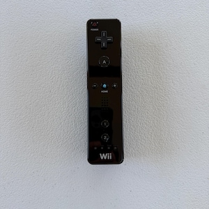 May include: A black rectangular video game remote controller featuring a power button, a directional pad, an A button, a home button, plus and minus buttons, buttons labeled 1 and 2, and the Wii logo printed in white at the bottom.