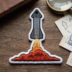 May include: An embroidered patch featuring a dark gray rocket ship launching upward. The rocket is surrounded by a white border and emits a plume of yellow and orange flames at the base, creating a cloud-like effect against the dark gray body.