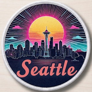 May include: A circular embroidered patch featuring a stylized Seattle skyline silhouette against a vibrant yellow and pink sunburst background. The word Seattle is printed in a large, light pink script font at the bottom, framed by a white border.