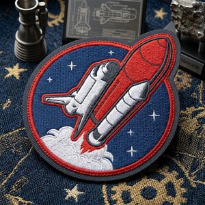 May include: A circular embroidered patch featuring a space shuttle and a red rocket booster against a dark blue background with white stars. The design includes a white exhaust plume at the base, all enclosed within a thick red circular border.