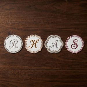 May include: Four embroidered coasters with elegant script letters: R, H, A, and S. Each coaster features a white center with a different colored border and letter. The coasters are arranged on a dark wood surface.