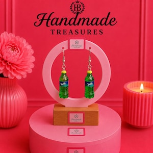May include: A pair of dangle earrings featuring miniature green plastic soda bottles with yellow caps and blue labels that read Sprite. The earrings are displayed on a circular pink stand with a wooden base, branded with the text Handmade Treasures.