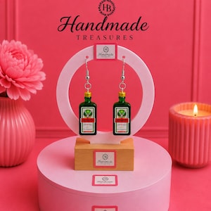 May include: A pair of dangle earrings featuring miniature green Jägermeister bottle charms with silver-toned metal hooks. The earrings are displayed on a circular white stand with a wooden base, featuring the text Handmade Treasures.