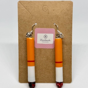 May include: A pair of novelty earrings shaped like cigarettes. Each earring has a white, orange, and red design, with a silver hook. The earrings are displayed on a brown card with a pink label that reads "Handmade Treasures."