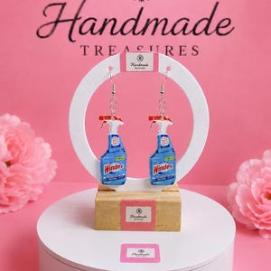 May include: A pair of dangle earrings featuring miniature blue Windex spray bottle charms with red triggers and white nozzles. The earrings hang from silver-toned metal hooks and are displayed on a white circular card labelled Handmade Treasures.