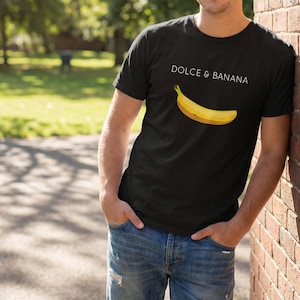 Peut inclure: T-shirt noir avec l'inscription "DOLCE & BANANA" et une illustration de banane jaune. Le t-shirt est porté avec un jean bleu. La personne est appuyée contre un mur de briques.