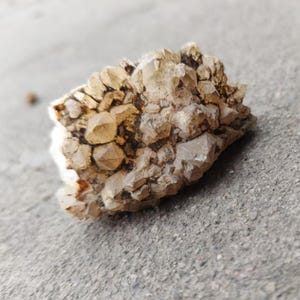 Op de afbeelding: Een cluster van kwartskristallen met een lichtbruine en heldere kleur. De kristallen hebben een ruw, getextureerd oppervlak en zijn samengeklonterd in een natuurlijke formatie. De kwarts staat tegen een grijze achtergrond.