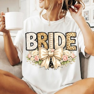 Könnte beinhalten: Weißes T-Shirt mit dem Wort "BRIDE" in einem dekorativen Design. Die Buchstaben haben unterschiedliche Muster, darunter goldener Glitzer, Perlen und Leopardenmuster. Eine große Schleife und ein Blumenarrangement befinden sich darunter.