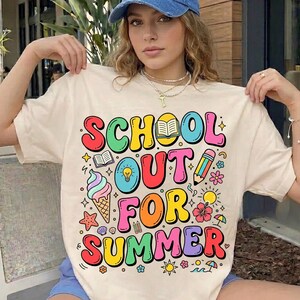 Può includere: T-shirt color crema con la scritta "SCHOOL OUT FOR SUMMER" in lettere colorate. Il design include illustrazioni di un gelato, una matita, fiori e un sole. Una donna indossa la maglietta.