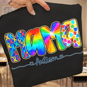 Puede incluir: Camiseta negra con la palabra "MAMA" en letras grandes y coloridas. Las letras están rellenas de arcoíris, piezas de rompecabezas y patrones de cintas. Debajo de la palabra "MAMA" está la palabra "Autismo" en cursiva.