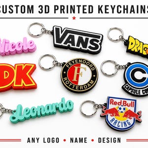 Op de afbeelding: Een verzameling op maat gemaakte 3D-geprinte sleutelhangers. Sleutelhangers met de namen "Nicole" en "Leonardo", het "Vans"-logo, "Dragon Ball", "DK", "Feyenoord Rotterdam", "Capsule Corp" en het Red Bull Racing-logo. De tekst luidt "ANY LOGO • NAME • DESIGN".