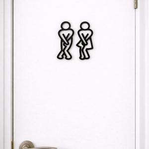 Op de afbeelding: Zwarte minimalistische toiletborden op een witte deur. De borden tonen gestileerde figuren van een persoon en een persoon, elk met gekruiste benen. Een zilveren deurklink is zichtbaar.