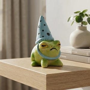 Könnte beinhalten: Eine kleine grüne Froschfigur aus Keramik mit einem hellblauen, spitz zulaufenden Hut mit dunkelblauen Punkten. Der Frosch hat einen neutralen, leicht nach unten gezogenen Mund und schmale Augen und steht auf einer hellen Holzoberfläche.