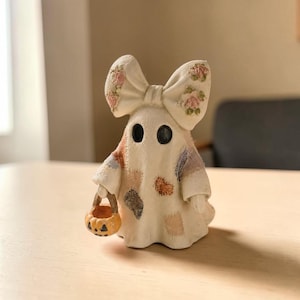 Op de afbeelding: Een klein gebroken wit spookbeeldje met een grote strik op het hoofd, versierd met roze bloemmotieven. Het spookje heeft zwarte ronde ogen, een textuurrijk patchworkontwerp op het lichaam en houdt een klein oranje pompoenemmertje vast.