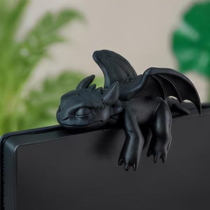 May include: A matte black dragon figurine with folded wings and closed eyes, designed to perch on the top edge of a flat screen monitor. The creature features pointed ears, a smooth surface, and a resting posture with its front limbs hanging down.