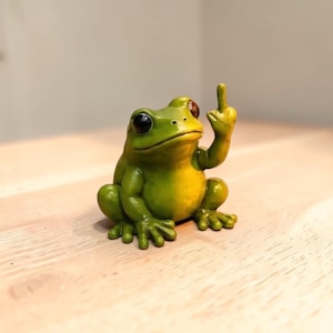Pode incluir: Uma pequena estatueta de sapo verde com grandes olhos pretos e uma textura lisa. O sapo é representado numa posição sentada com um dos membros anteriores levantado, estendendo o dedo médio para cima num gesto dirigido ao observador.