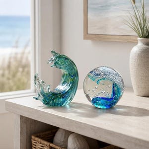 Könnte beinhalten: Zwei dekorative Glasobjekte mit ozeaninspirierten Designs. Eines ist eine geschwungene, wellenförmige Skulptur in Blau- und Grüntönen. Das andere ist eine klare Glaskugel, die ein blau-grünes Wellenmuster mit eingeschlossenen Luftblasen enthält.
