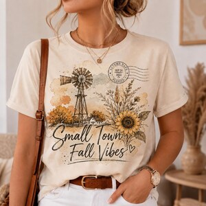 May include: A cream-colored short-sleeve t-shirt featuring a graphic print of a windmill, a sunflower, and the text "Small Town Fall Vibes" with a postmark stamp dated SEP 24 2024. The design includes watercolor-style orange and yellow accents.