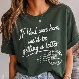 May include: Dark green t-shirt with white text that reads "If Paul were here, we'd be getting a letter." A postal service stamp graphic is below the text. The shirt has rolled-up sleeves.