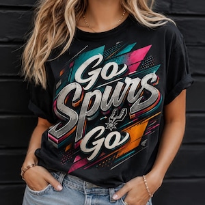 Op de afbeelding: Zwart t-shirt met de tekst "Go Spurs Go" in een 3D-ontwerp met roze, turquoise en oranje accenten. Het shirt toont het Spurs-logo en een geometrische achtergrond.