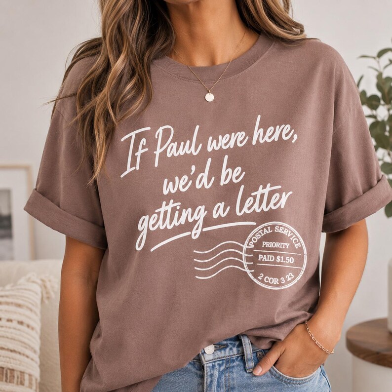 May include: A brown t-shirt with rolled-up sleeves featuring white text that reads "If Paul were here, we'd be getting a letter." Below the text is a postal service stamp graphic.