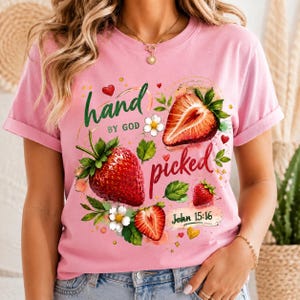 Puede incluir: Camiseta rosa con un diseño de fresas y el texto "hand picked by God" y "John 15:16". El diseño incluye fresas rojas, hojas verdes y flores blancas. La camiseta tiene mangas cortas.