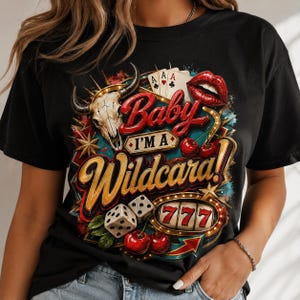 Op de afbeelding: Zwart T-shirt met een kleurrijk grafisch ontwerp. Het ontwerp bevat een schedel van een stier, speelkaarten, dobbelstenen, kersen en de tekst "Baby I'm a Wildcard!" in een vintage casinostijl.