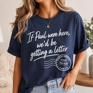 May include: Navy blue t-shirt with the text "If Paul were here, we'd be getting a letter" in white script. A postal service stamp graphic is below the text. The shirt has rolled-up sleeves.