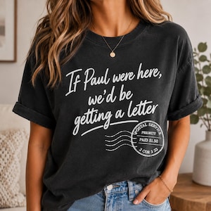 May include: Black t-shirt with the text "If Paul were here, we'd be getting a letter" in white script. A postal service stamp graphic is below the text. The shirt has rolled-up sleeves and is worn by a person.