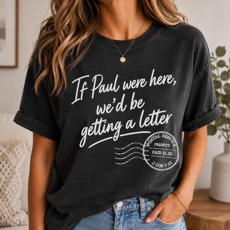 May include: Black t-shirt with the text "If Paul were here, we'd be getting a letter" in white script. A postal service stamp graphic is below the text. The shirt has rolled-up sleeves and is worn by a person.